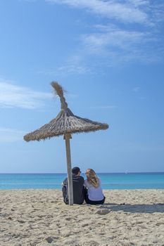 Young natural and casual sporty couple sit on beach under parasol and watch the ocean horizon in Mallorca, Spain.