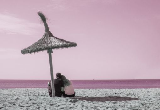 Young couple sit closely together on beach under parasol and watch the ocean horizon in surrealistic romantic pink