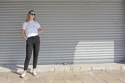 Full body photo of young casual sporty dressed blonde woman with sunglasses in white t-shirt against corrugated iron wall street style