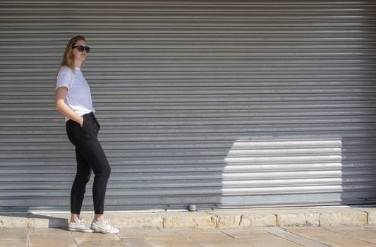 Full body photo of young casual sporty dressed blonde woman with sunglasses in white t-shirt against corrugated iron wall street style