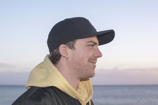 Profile shot of handsome smiling natural and casual looking male in late twenties with hood jacket and cap looks straight forward, ocean horizon behind