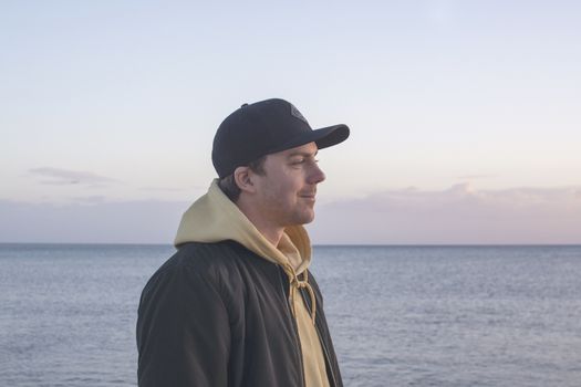 Profile shot of handsome smiling natural and casual looking male in late twenties with hood jacket and cap looks straight forward, ocean horizon behind