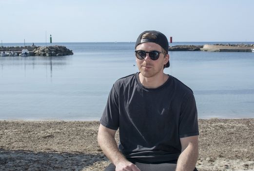 Young handsome happy man enjoys the sun in black glasses cap backwards and black tee by the beach