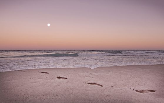 Empty sandy beach with footprints, ocean waves and full moon in expressive dusk orange and purple toned in color Living Coral.