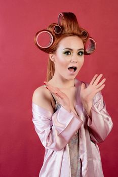 Young redhead woman with hair curlers dressed in peignoir, dressing gown posing on a burgundy background. Advertising concept with copyspace for design. Expression of different emotions. Pin-up style.