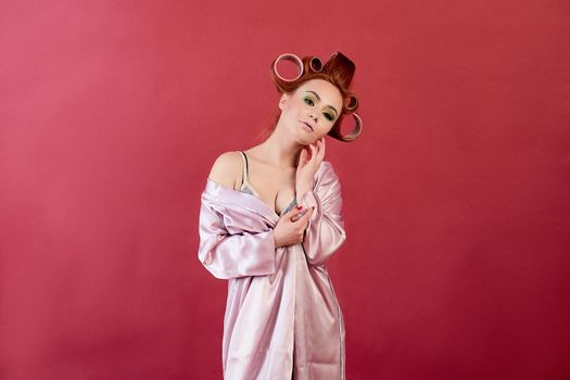 Young redhead woman with hair curlers dressed in peignoir, dressing gown posing on a burgundy background. Advertising concept with copyspace for design. Expression of different emotions. Pin-up style.