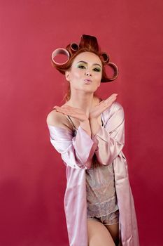 Young redhead woman with hair curlers dressed in peignoir, dressing gown posing on a burgundy background. Advertising concept with copyspace for design. Expression of different emotions. Pin-up style.