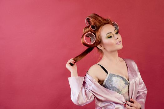 Young redhead woman with hair curlers dressed in peignoir, dressing gown posing on a burgundy background. Advertising concept with copyspace for design. Expression of different emotions. Pin-up style.