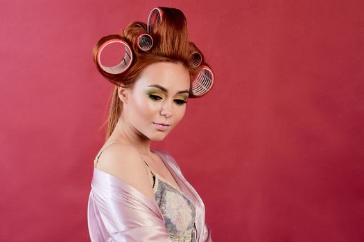 Young redhead woman with hair curlers dressed in peignoir, dressing gown posing on a burgundy background. Advertising concept with copyspace for design. Expression of different emotions. Pin-up style.