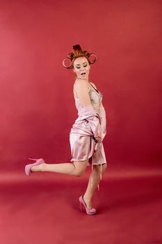 Young redhead woman with hair curlers dressed in peignoir, dressing gown posing on a burgundy background. Advertising concept with copyspace for design. Expression of different emotions. Pin-up style.