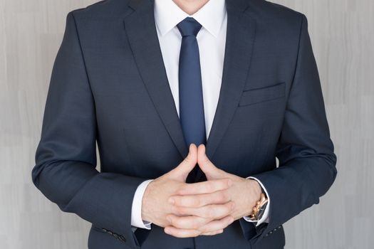 Torso of anonymous businessman standing with hands in lowered steeple, wearing beautiful fashionable classic navy blue suit against grey backgound.