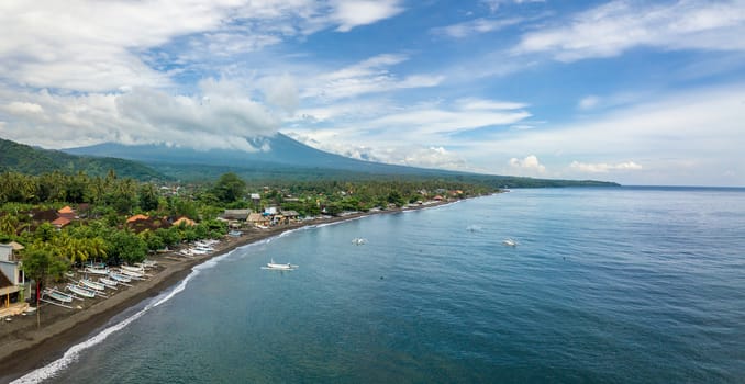 Panoramic aerial view of Amed beach in Bali, Indonesia. Traditional fishing boats called jukung on the black sand beach and Mount Agung volcano in the background, partially covered by clouds.