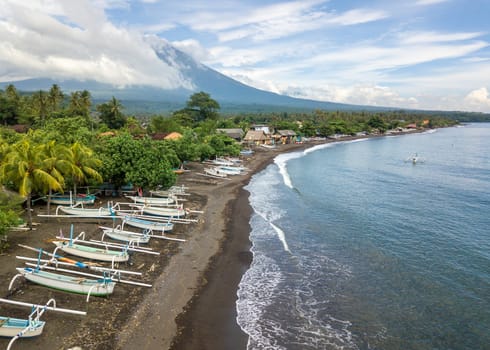 Aerial view of Amed beach in Bali, Indonesia. Traditional fishing boats called jukung on the black sand beach and Mount Agung volcano in the background, partially covered by clouds.