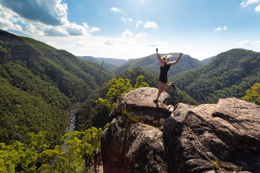 Spirited woman jumping on a high cliff ledge.  Motion in her actions.  Mountain valley views.
