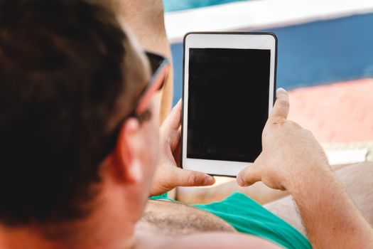 A young man in sunglasses holds a tablet in his hands. Freelancer job concept on the beach and vacation.
