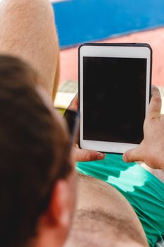 A young man in sunglasses holds a tablet in his hands. Freelancer job concept on the beach and vacation.