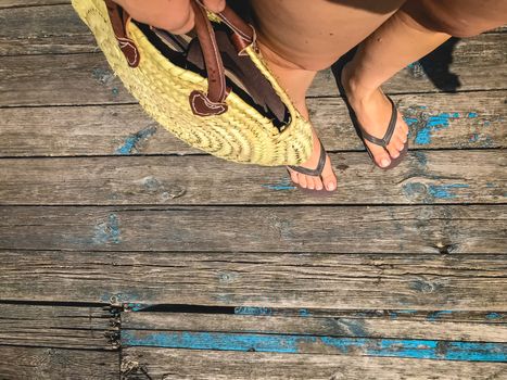 Top view, photo of legs in beach flip-flops and with a straw bag in hand on a wooden old floor. Photos on vacation, beach, summer.