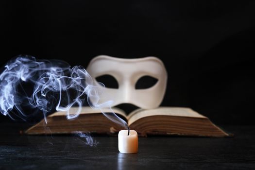 White classical Venetian mask on book near extinguished candle on dark background