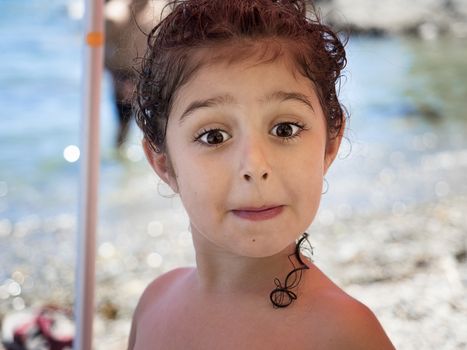 portrait of a kid mocking to the camera, she is shirtless on the beach, her curly hair is wet and she has it picked up, some curls hang down her neck, in the unfocused background you see the seashore