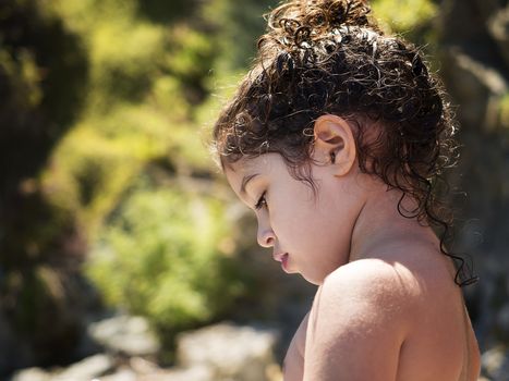 portrait of a kid looking down pensively, she is shirtless on the beach, her curly hair is wet and she has it picked up, some curls hang down her neck, the green vegetation background is out of focus