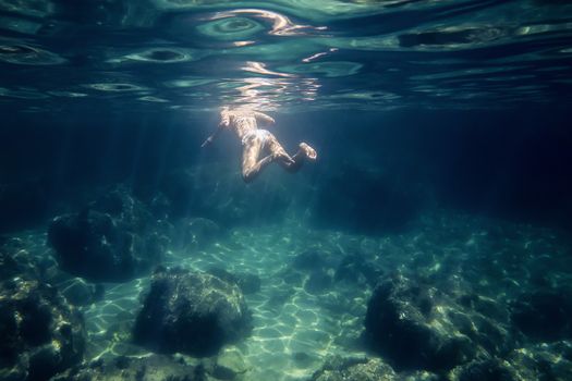 woman swimming in the sea seen from under water. The sun reflects on her back and on the curly surface of the sea, also illuminates the rocks of the bottom and light rays are formed in the water