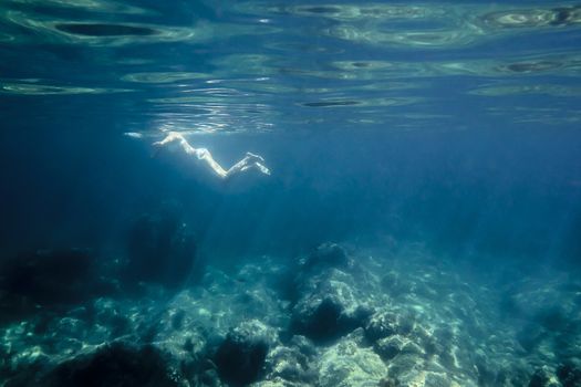 underwater view of a woman relaxing leisurely at sea. The sun reflects on her back and on the curly surface of the sea, also illuminates the rocks of the bottom and light rays are formed in the water