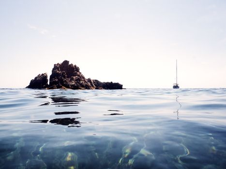 minimalist photograph of a typically Mediterranean marine landscape. The sky is clear, the surface of the sea is calm and there is a sailboat with folded sails near an islet of rocks