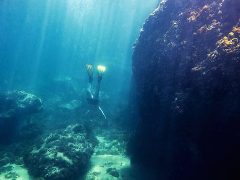 Underwater view of a man diving towards the marine depth with his yellow fins. Sun rays enter the water illuminating the huge rock next to it and the bottom of sand and stones