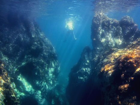 Man floats relaxed in the ocean between huge rocks that come from the bottom to the surface of the water. The view is from under the sea and the sunbeams enter in the water and illuminate the depths