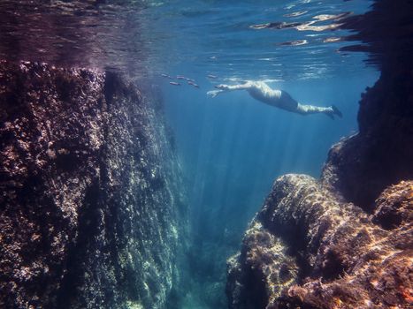 Underwater view of a man swimming placidly in the sea with some fish. Sun rays are drawn in the crystal clear water and illuminate the rocks and curly surface of the blue ocean