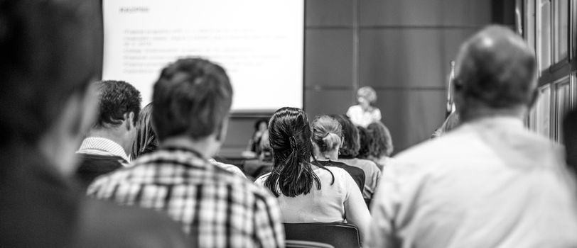 Business and entrepreneurship symposium. Female speaker giving talk at business meeting. Audience in conference hall. Rear view of unrecognized participant in audience. Black and white image.