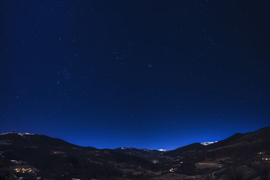 mountain landscape under night sky full of stars, you can see the lights of the houses in the valley and some snowy mountains