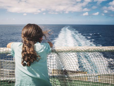 little girl leaning on the railing looks at the wake left by the cruise ship in which she travels, the wind moves her hair and the white foam of the boat moves through the blue ocean. copy space