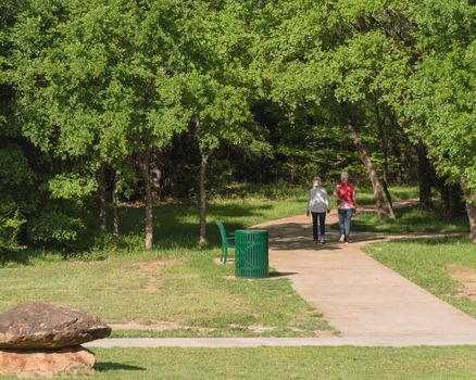 Rear view of senior Caucasian female couple walking in the park. Nature recreation area with trail, trees, bench, trash bin and decorative landscape rock, boulder. Happy retirement concept