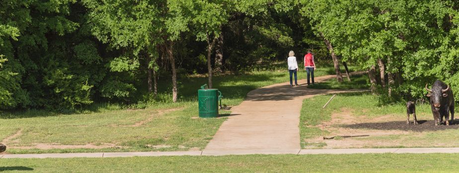 Panorama rear view of senior Caucasian female couple walking in the park. Nature recreation area with trail, trees, bench, trash bin and decorative landscape rock, boulder. Happy retirement concept