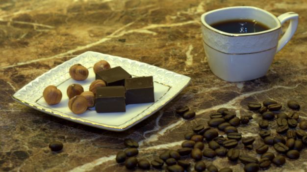 A glass cup of black coffee and a saucer with chocolates on a wooden table. Close-up shot.