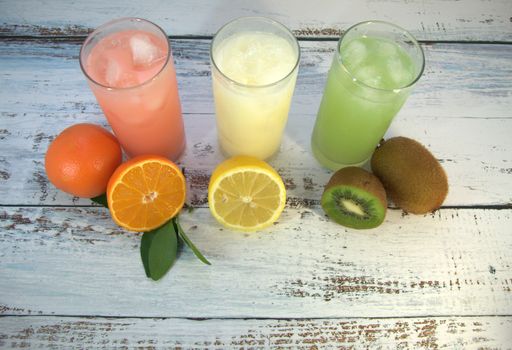 Three glass glasses with a refreshing juice and ice, on a textile stand, whole and sliced half of an orange with leaves, lemon and kiwi, lay on a white wooden table. Close-up.