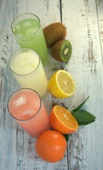 Three glass glasses with a refreshing juice and ice, on a textile stand, whole and sliced half of an orange with leaves, lemon and kiwi, lay on a white wooden table. Close-up.