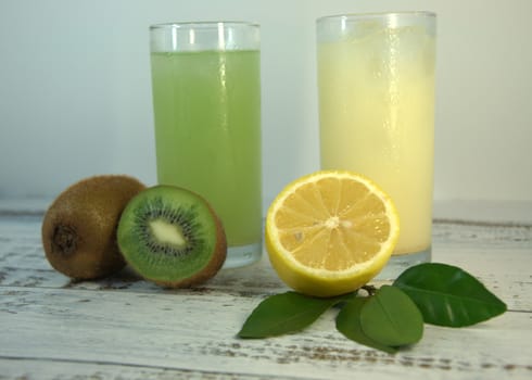 Two glass glasses with a refreshing juice and ice, on a textile stand, whole and sliced half of an orange with leaves and kiwi, lies on a white wooden table. Close-up.