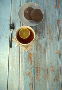 A cup of tea with lemon, a spoon and two chocolate cakes on a plate lie on a wooden table. Close-up.