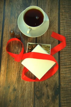 A cup of tea, a white box of chocolates with a red satin ribbon on a wooden table. Close-up.