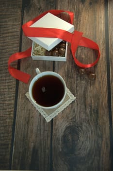 A cup of tea, a white box of chocolates with a red satin ribbon on a wooden table. Close-up.