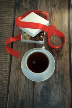 A cup of tea, a white box of chocolates with a red satin ribbon on a wooden table. Close-up.