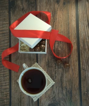 A cup of tea, a white box of chocolates with a red satin ribbon on a wooden table. Close-up.