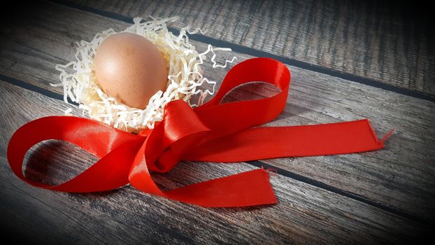 Chicken egg in a nest of straw and red satin ribbon on a wooden table. Close-up.