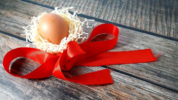 Chicken egg in a nest of straw and red satin ribbon on a wooden table. Close-up.