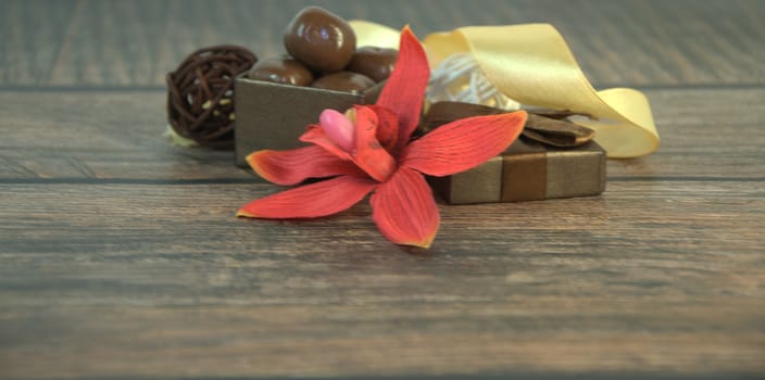 A box of chocolates, decorative balls, a bud of red orchid and beige ribbon on a wooden table. Close-up.