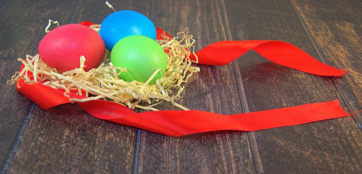 Painted Easter eggs in a nest of straw on a scarlet ribbon lie on a wooden table. Close-up.