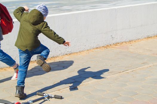 Teen boy in green jacket and jeans falls off his scooter on outdoor in Park.