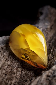 A piece of yellow semi opaque natural amber, classification color Bastard, has cracks inside. Polished, has a bead shape. Placed on dark stoned wood texture.